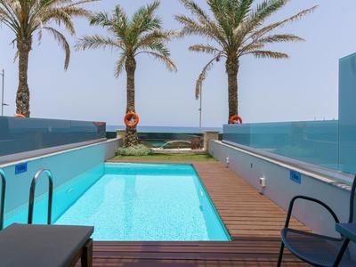 Área de piscina moderna con agua azul, terraza de madera, sillas y palmeras al fondo.