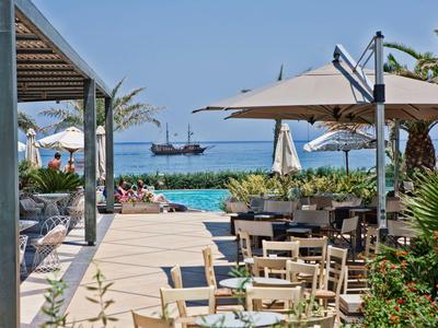 Restauracja przy plaży z drewnianymi stołami i parasolami, widok na morze i żaglowiec.