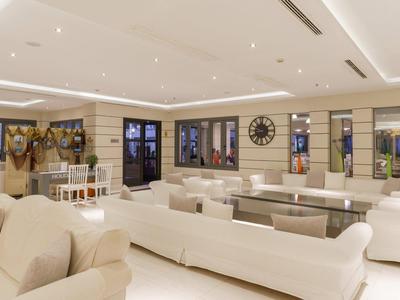 Ruime hotel lounge met beige banken, glazen tafels, grote klok en heldere verlichting.