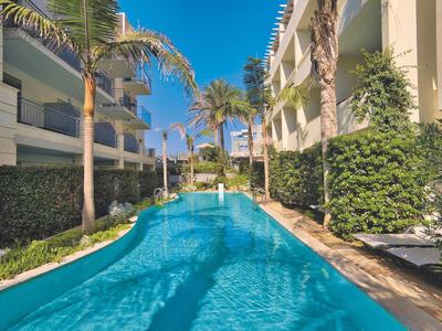 Schmaler Pool zwischen hellen Gebäuden, umgeben von Palmen und grünen Hecken, unter blauem Himmel.