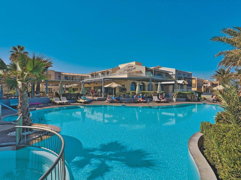 Helderblauw zwembad met palmbomen en een hotelgebouw onder een zonnige hemel.