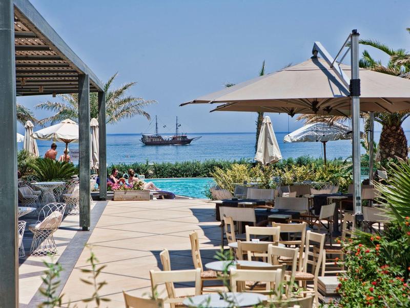 Strandresort met zitplaatsen buiten, parasols, palmbomen en een schip op zee.