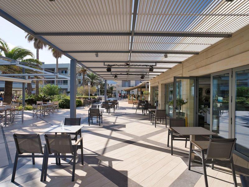 Buitenterras van hotel met tafels en stoelen onder een overdekte luifel omringd door palmbomen.