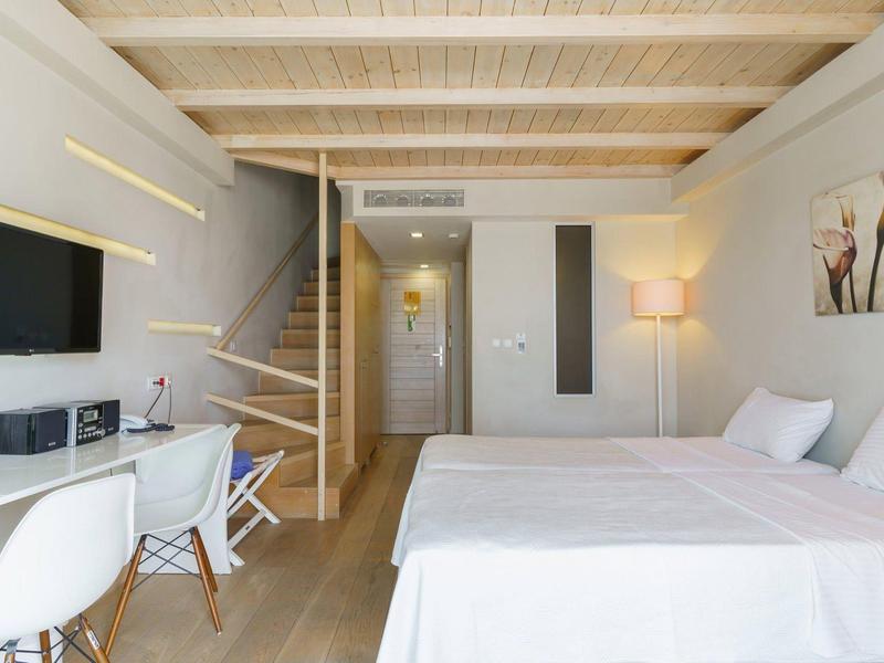 Lichte hotelkamer met groot bed, houten plafondbalken, tv, bureau en trap.