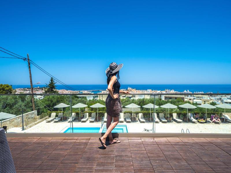 Frau schaut von Terrasse auf Pool, Sonnenschirme und Aussicht aufs Meer.