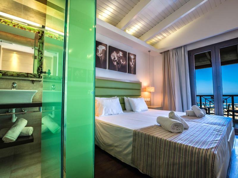 Modernes Hotelzimmer mit Doppelbett, Glasdusche und Meerblick vom Balkon bei Dämmerung.