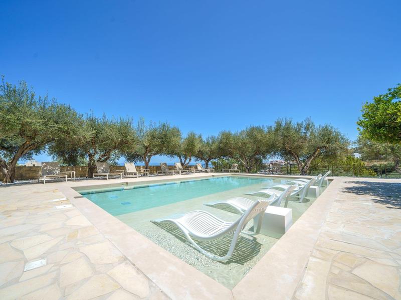 Kleiner Pool mit Liegestühlen auf Steinboden und Bäumen im Hintergrund unter blauem Himmel.