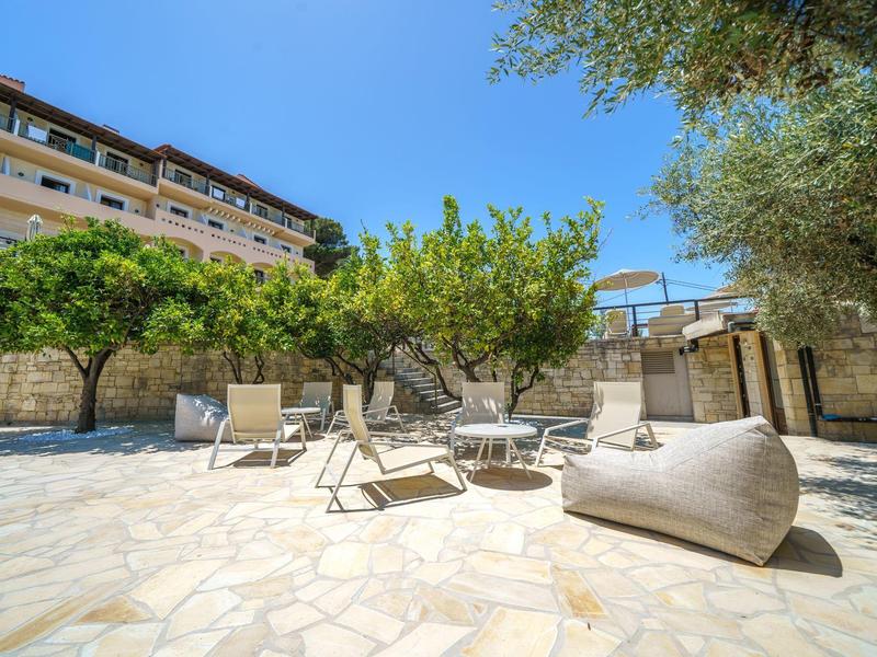 Sonnige Terrasse mit Liegestühlen, Tisch und Bäumen neben einem Hotelgebäude.