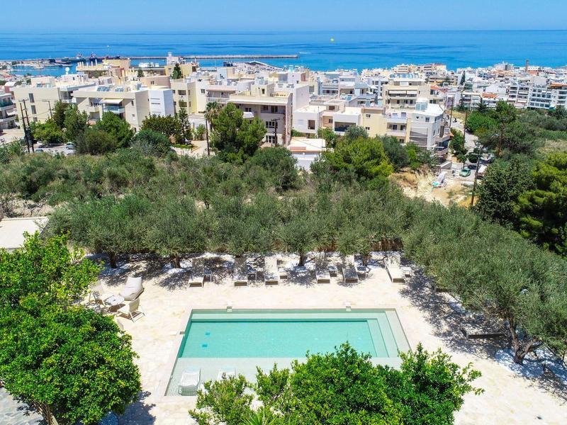 Blick auf einen Pool umgeben von Bäumen mit weißen Gebäuden und blauem Meer im Hintergrund.