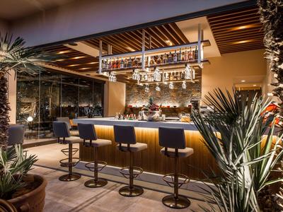 Moderne Bar mit Holzdecke, schwarzen Barhockern und Pflanzen in Töpfen, warm beleuchtet.