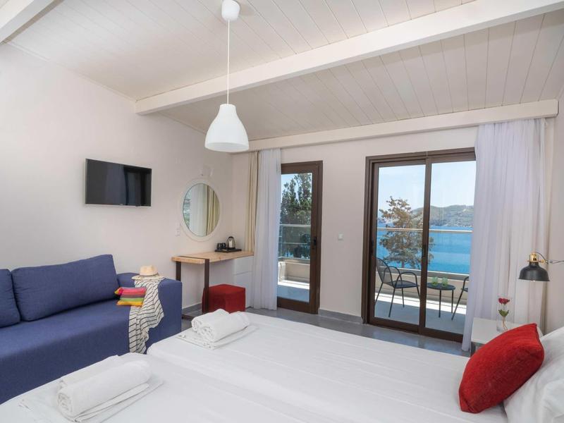 Světý hotelový pokoj s manželskou postelí, pohovkou a výhledem na moře přes dvojité skleněné dveře.