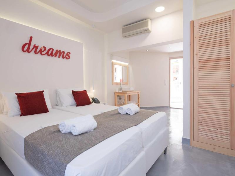 Světle zařízený hotelový pokoj s velkou postelí, šedou přikrývkou a červenými doplňky.