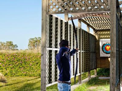 Młody mężczyzna stoi pod drewnianą pergolą z tarczami, otoczony trawą i czystym niebem.