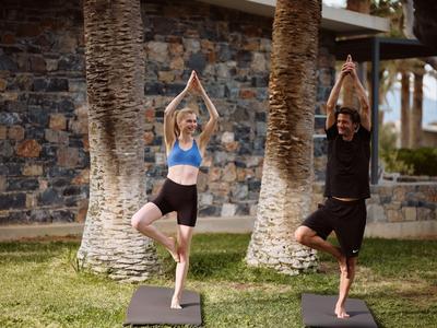 Zwei Personen praktizieren Yoga im Garten vor einer Steinmauer und Palmenstämmen.