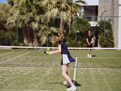 Zwei Personen spielen Tennis auf einem Rasenplatz neben einem Hotelgebäude mit Palmen.