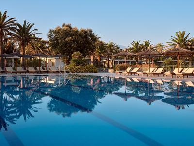 Großer Hotelpool mit Sonnenliegen und Palmen bei klarem Himmel am späten Nachmittag.