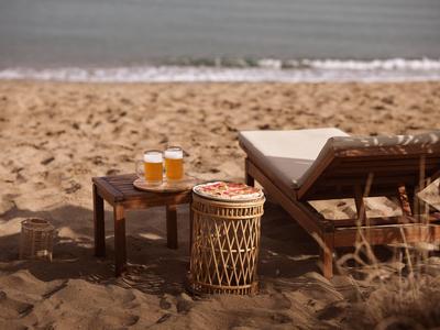 Zwei Gläser Bier und eine Pizza neben Liegestuhl am Sandstrand mit Blick aufs Meer.