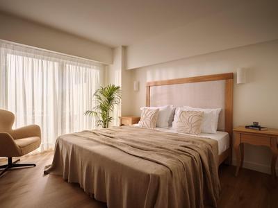 Helles Hotelzimmer mit großem Bett, beiger Bettwäsche, Sesseln und seitlichem Nachttisch.