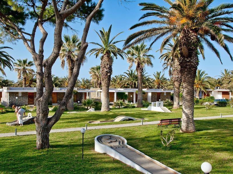 Zielony ogród hotelowy z palmami, polem do minigolfa i białymi bungalowami w tle.