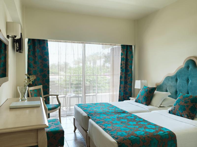 Prosto umeblowany pokój hotelowy z podwójnym łóżkiem, biurkiem i balkonem z widokiem na zieleń.