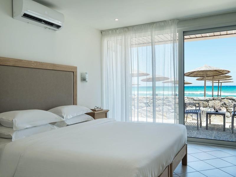 Jasny pokój hotelowy z podwójnym łóżkiem i widokiem na taras z leżakami i morze.