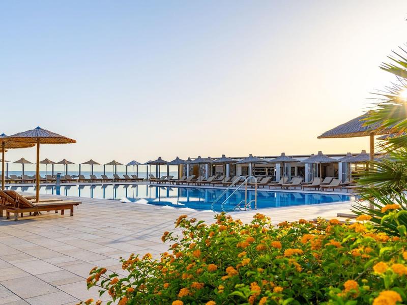 Basen na zewnątrz hotelu z leżakami, parasolami i kwiatami na pierwszym planie o zachodzie słońca.