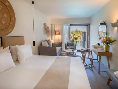 Chambre d'hôtel lumineuse avec grand lit, coin salon, bureau et vue sur une terrasse.