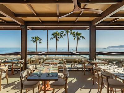 Offenes Restaurant mit Holzdecke und Blick auf Palmen und Meer am Strand.