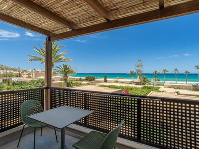 Balkon mit Tisch und Stühlen bietet Blick auf Strand, Palmen und Meer unter blauem Himmel.