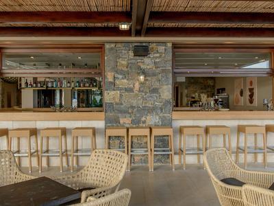 Moderner Hotelbarbereich mit Steintresen, Holzhockern und geflochtenen Stühlen unter Holzbalkendecke.