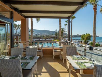 Terrasse mit gedeckten Tischen, Blick auf Pool, Meer und Berge bei klarem Himmel.