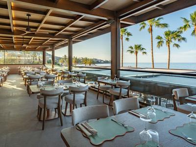 Terrasse eines Hotels mit gedeckten Tischen, Blick auf Meer und Palmen bei Sonnenuntergang.