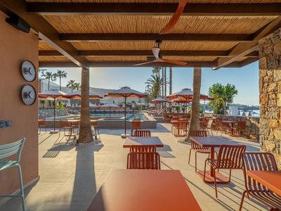 Überdachte Terrasse mit Tischen und Sesseln blickt auf Pool und Palmen in Hotelanlage.