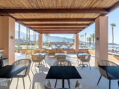 Überdachte Terrasse mit Tischen und Stühlen mit Blick auf Palmen und Meer.