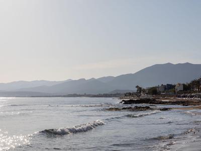 Küstenlinie mit sanften Wellen, felsigem Strand und Bergen im Hintergrund bei klarem Himmel.