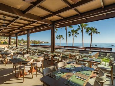 Überdachte Terrasse eines Hotels mit Tischen und Stühlen, Blick auf Palmen und Meer.