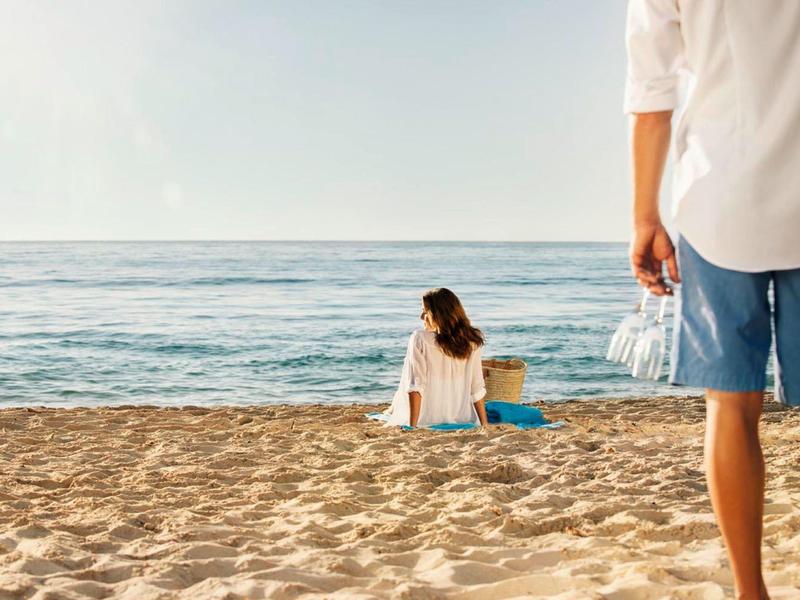 Femme assise sur la plage près de l'océan, homme tenant des bouteilles d'eau debout à proximité sous un ciel clair.