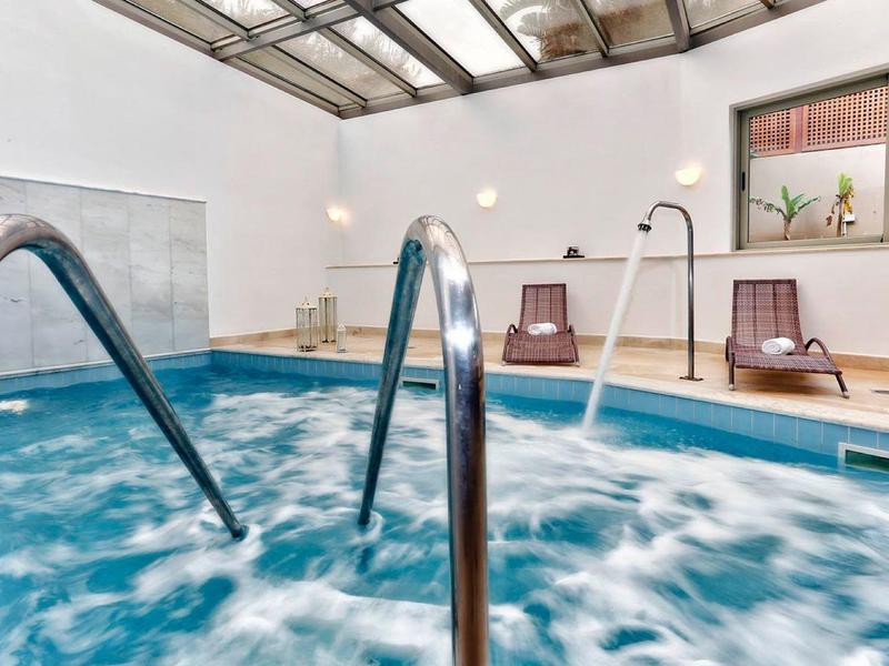 Piscine intérieure avec eau bleue, rampes métalliques et chaises longues près de grandes fenêtres.