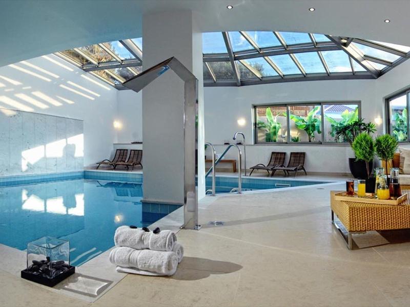 Piscine intérieure lumineuse avec plafond en verre, chaises longues, plantes et sièges confortables.