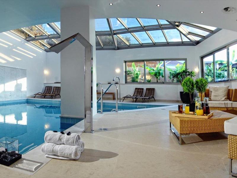 Espace piscine intérieure avec plafond en verre ensoleillé, chaises longues et coin salon moderne.