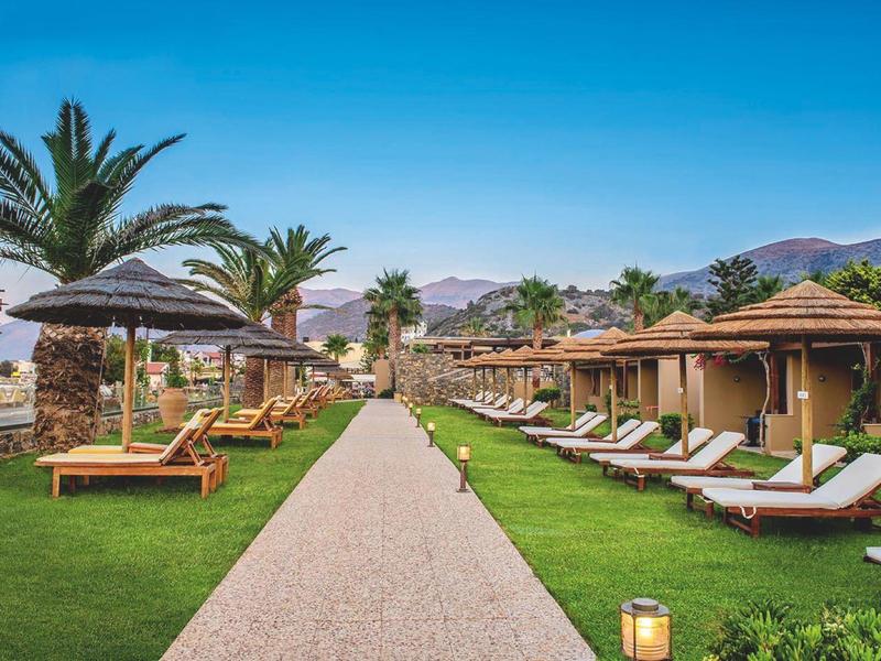 Chemin entre pelouse et transats menant à des bungalows en bord de mer avec palmiers et montagnes.