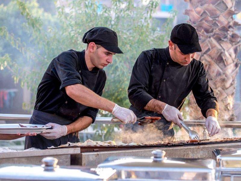 Deux chefs grillent des brochettes en plein air près des palmiers, préparant la nourriture au soleil.