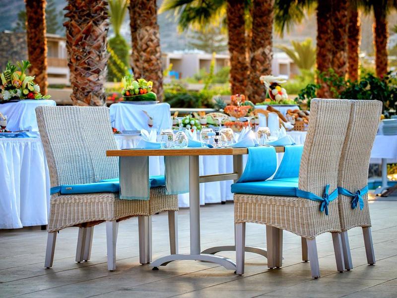 Table de repas extérieure avec chaises en osier attachées avec des rubans bleus sous des palmiers.