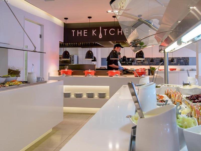 Espace buffet moderne avec une variété de plats exposés et des chefs préparant des repas dans une cuisine lumineuse.