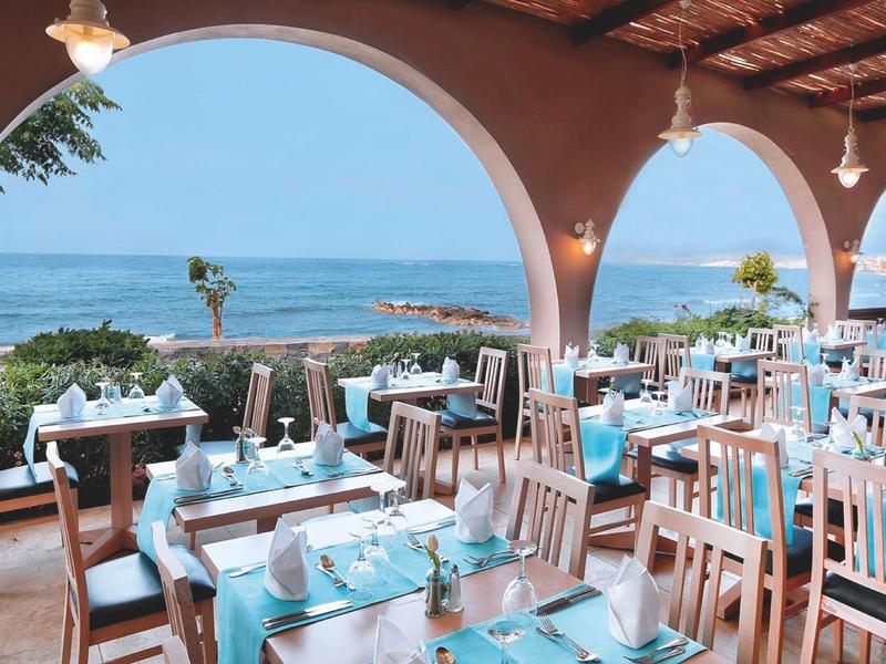 Restaurant en plein air avec vue sur l'océan, chaises en bois et tables avec nappes bleues.