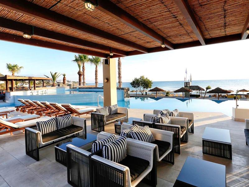 Espace lounge extérieur couvert avec canapés donnant sur une piscine et transats sous parasols.