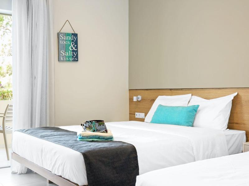 Chambre d'hôtel avec un lit, draps blancs, coussin turquoise et une fenêtre avec des rideaux légers.