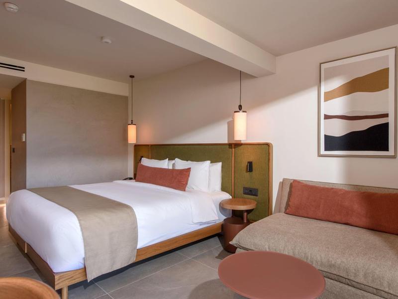 Chambre d'hôtel moderne avec lit, canapé, table ronde, suspensions et art mural abstrait.