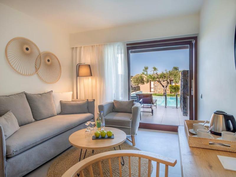 Salon lumineux avec canapé gris, fauteuils, meubles en bois, vue sur terrasse avec piscine et palmiers.
