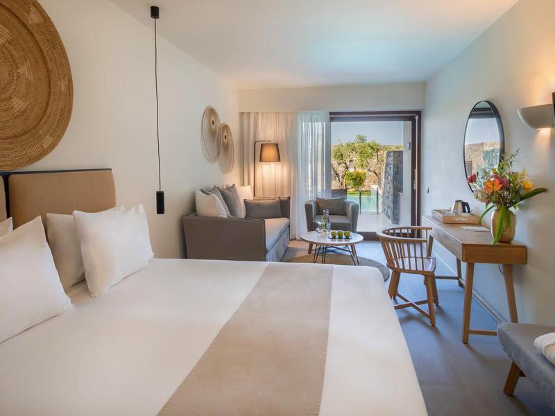 Chambre d'hôtel lumineuse avec grand lit, coin salon, bureau et vue sur une terrasse.